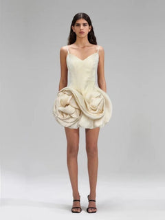 Woman wearing a unique cream-colored dress with a large floral embellishment on a plain background