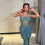 Woman wearing an aqua crystal-embellished sheer gown with shimmering sequins, posing confidently indoors with a mirror in the background