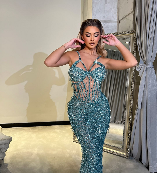 Woman wearing an aqua crystal-embellished sheer gown with shimmering sequins, posing confidently indoors with a mirror in the background