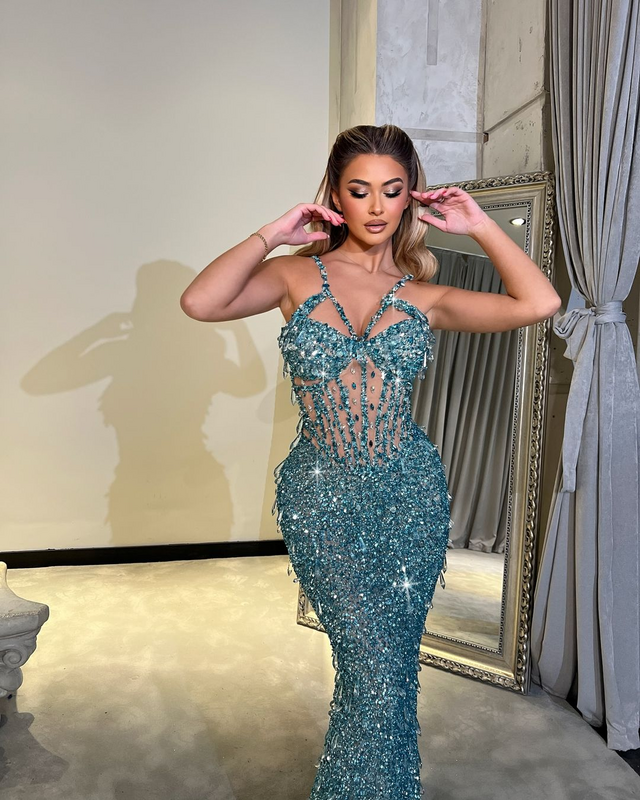 Woman wearing an aqua crystal-embellished sheer gown with shimmering sequins, posing confidently indoors with a mirror in the background