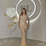 Woman in champagne embellished long-sleeve gown standing under soft lighting, beside large paper flowers