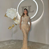 Woman in champagne embellished long-sleeve gown standing under soft lighting, beside large paper flowers