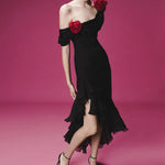 Woman in a romantic off-shoulder black dress with red rose accents and layered ruffles, standing against a pink background