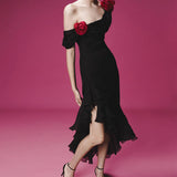 Woman in a romantic off-shoulder black dress with red rose accents and layered ruffles, standing against a pink background