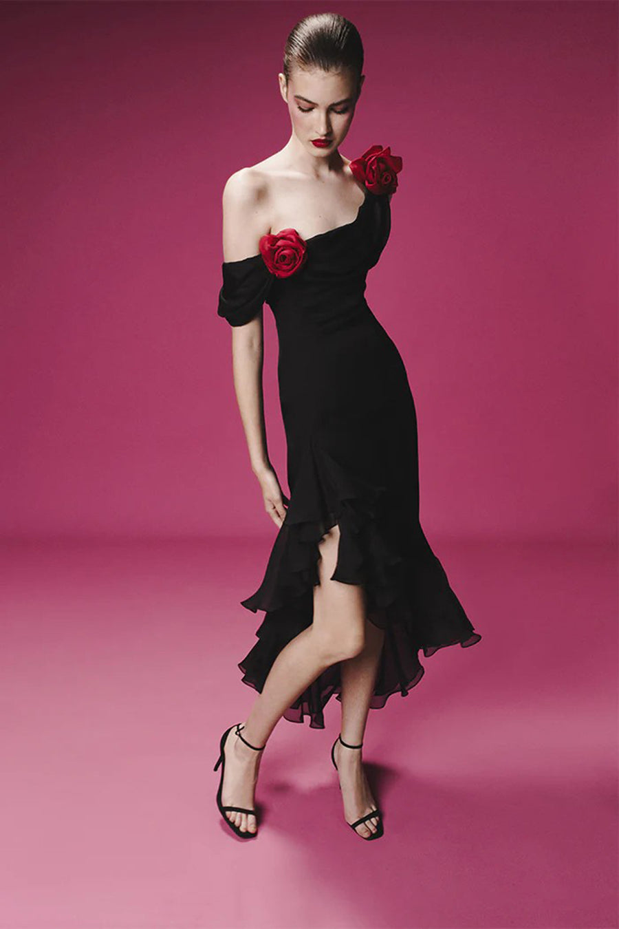 Woman in a romantic off-shoulder black dress with red rose accents and layered ruffles, standing against a pink background